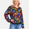 Eine blonde Frau trägt einen hochwertigen, farbenfrohen Patchwork-Pullover aus Alpaka und Jeans vor einem weißen Hintergrund. Der Pullover hat ein einzigartiges Design aus quadratischen Mustern in verschiedenen Farben.