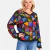 Eine blonde Frau trägt einen hochwertigen, farbenfrohen Patchwork-Pullover aus Alpaka und Jeans vor einem weißen Hintergrund. Der Pullover hat ein einzigartiges Design aus quadratischen Mustern in verschiedenen Farben.
