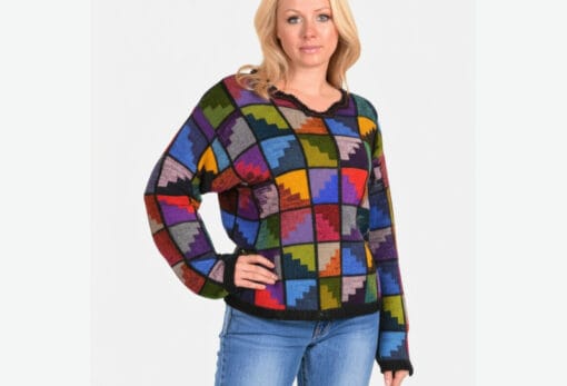 Eine blonde Frau trägt einen hochwertigen, farbenfrohen Patchwork-Pullover aus Alpaka und Jeans vor einem weißen Hintergrund. Der Pullover hat ein einzigartiges Design aus quadratischen Mustern in verschiedenen Farben.